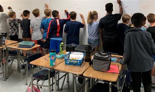 Students in class standing at a white large wall white board completing math problems. 